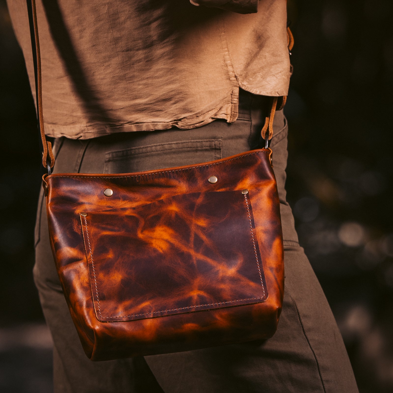 A person wearing a brown shirt and dark pants carries a brown leather shoulder bag with a marbled orange-brown pattern. The bag has a front pocket and metal snap details, photographed against a dark blurred outdoor background.