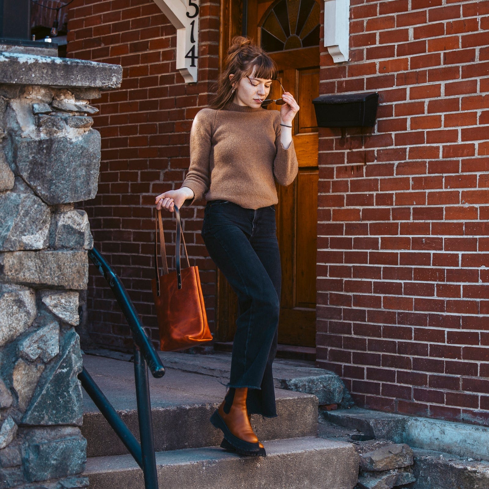 Woman carrying a Leather Everyday Tote in English Tan down brick steps outside a red-brick house.