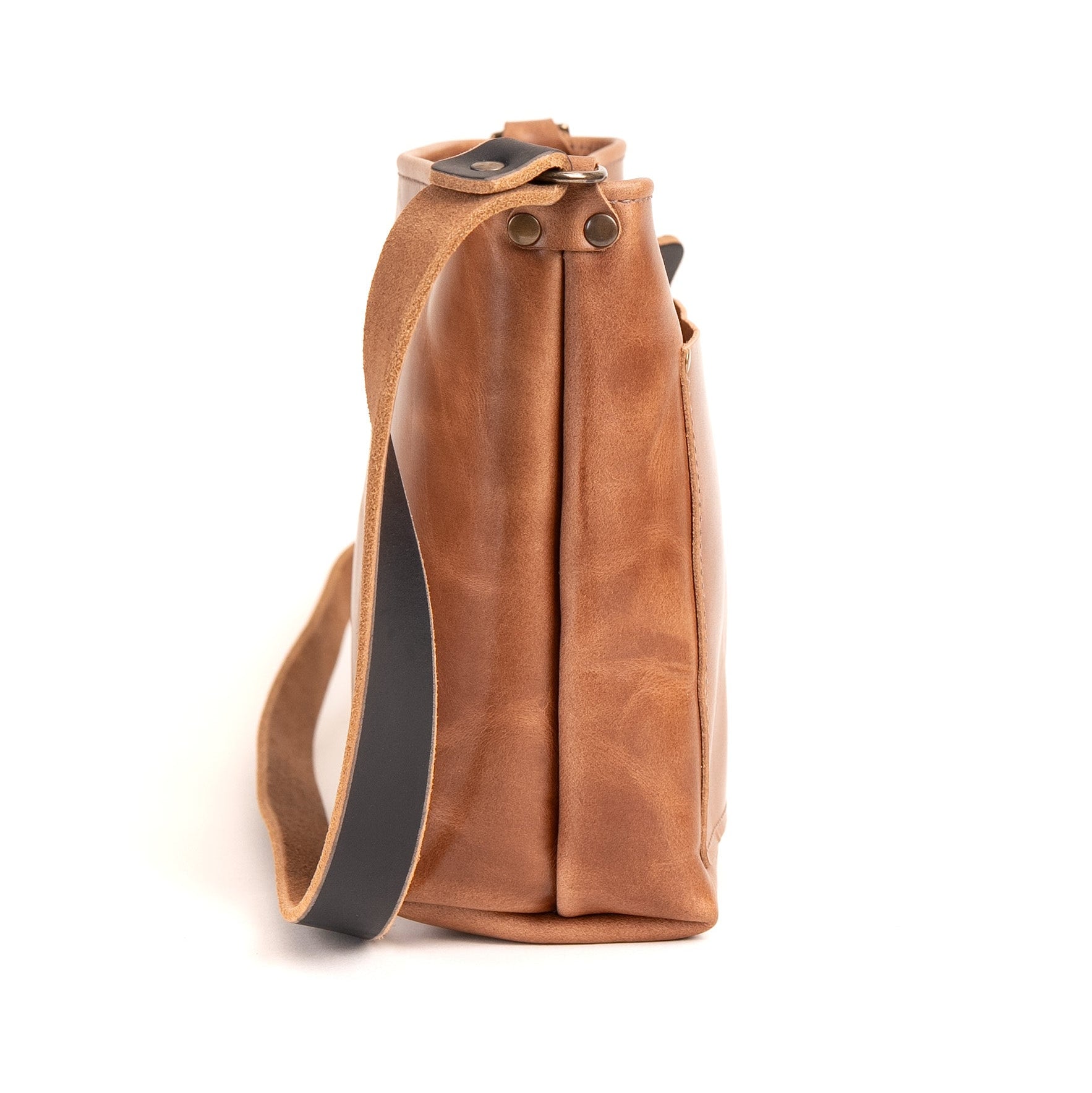 A side view of a tan leather shoulder bag with a black strap and brass rivets, shown on a white background.