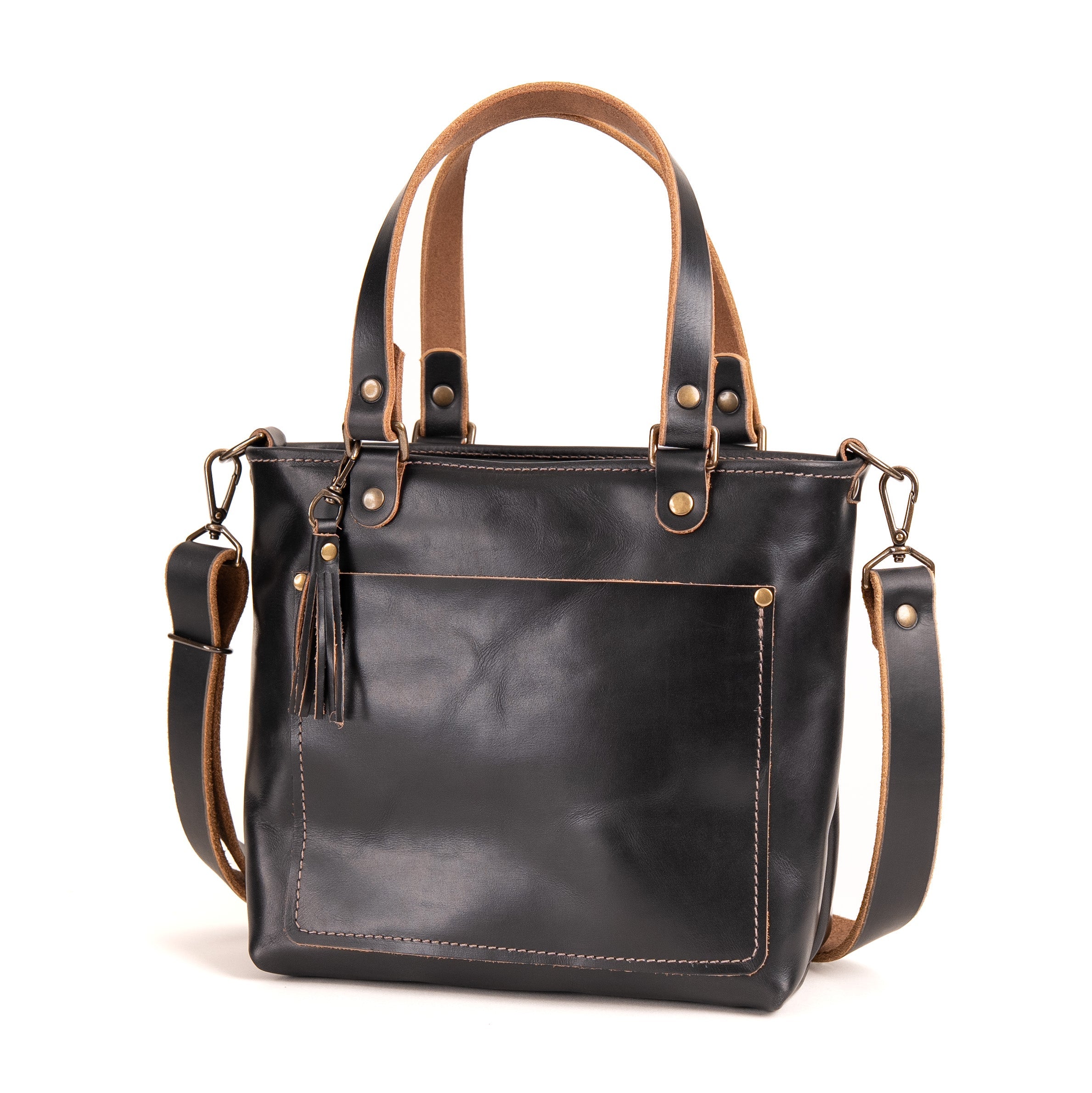 Black leather tote bag with tan handles, trim, and lining, featuring a front slip pocket with a leather tassel charm, gold-tone hardware, and an adjustable shoulder strap, shown on a white background.