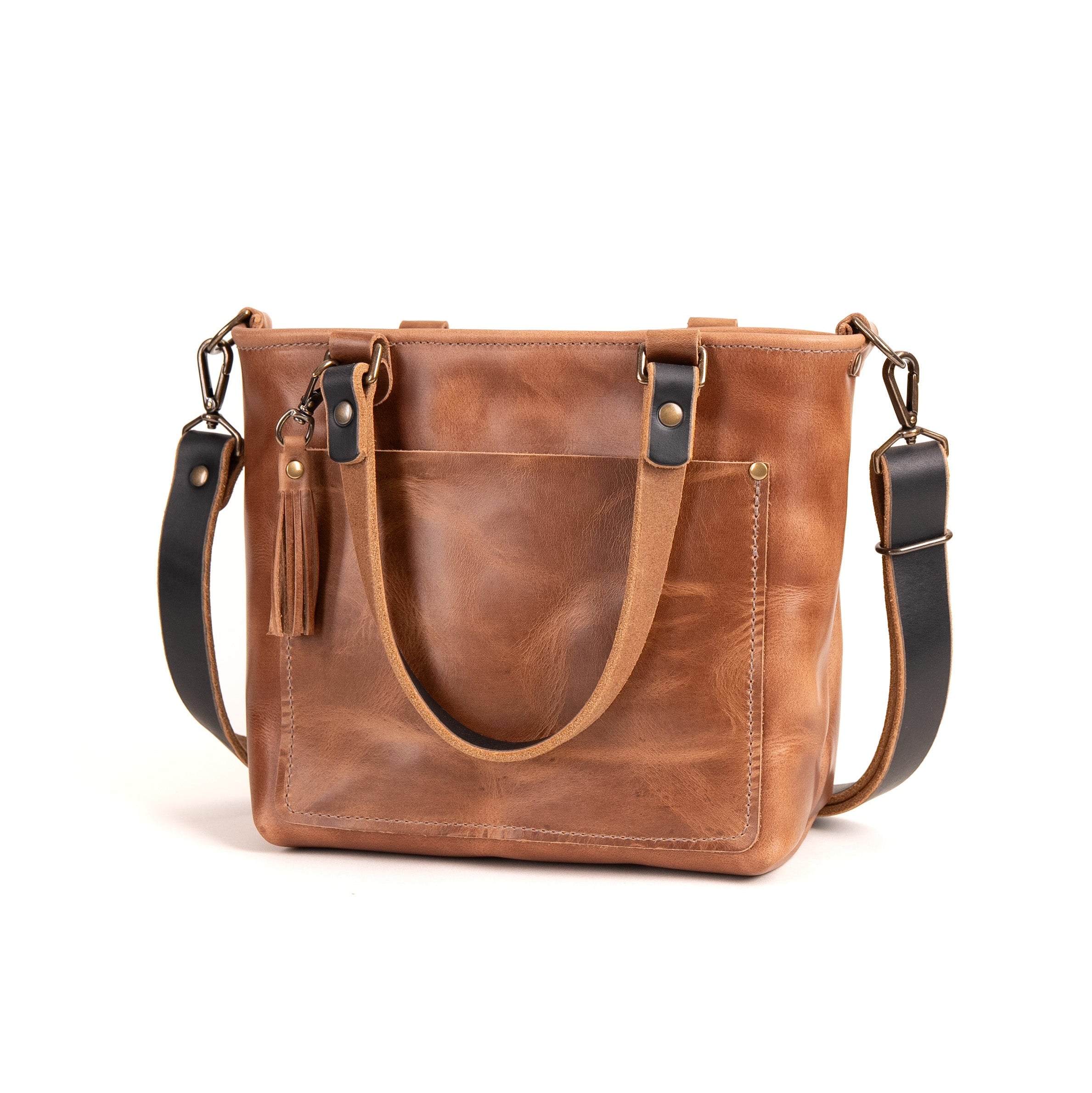A brown distressed leather tote bag with a front pocket, dual top handles, a tassel detail, and a detachable black shoulder strap, shown on a white background.