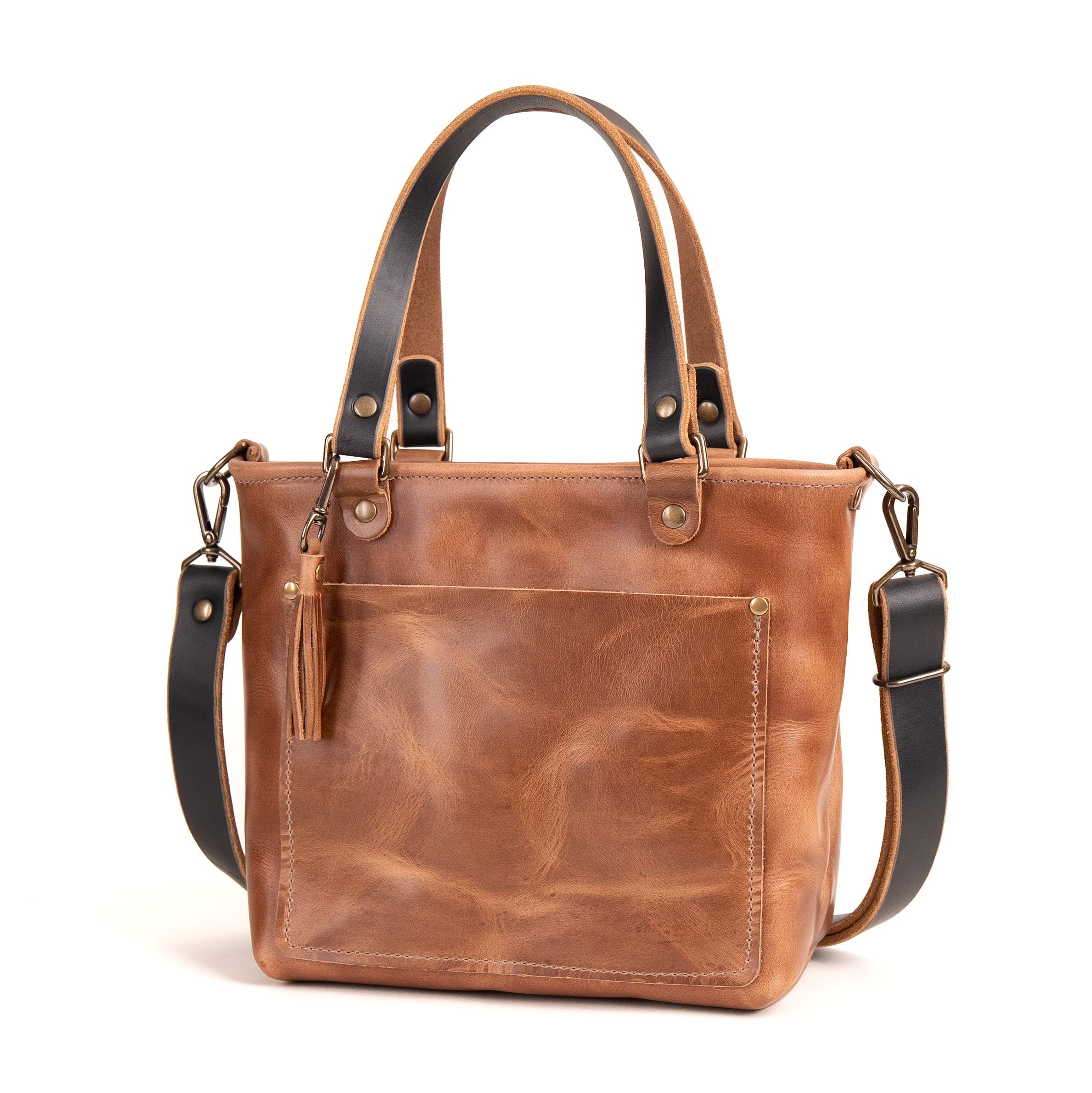 A brown leather tote bag with black leather handles and shoulder strap, featuring a front pocket and a tassel detail, shown on a white background.