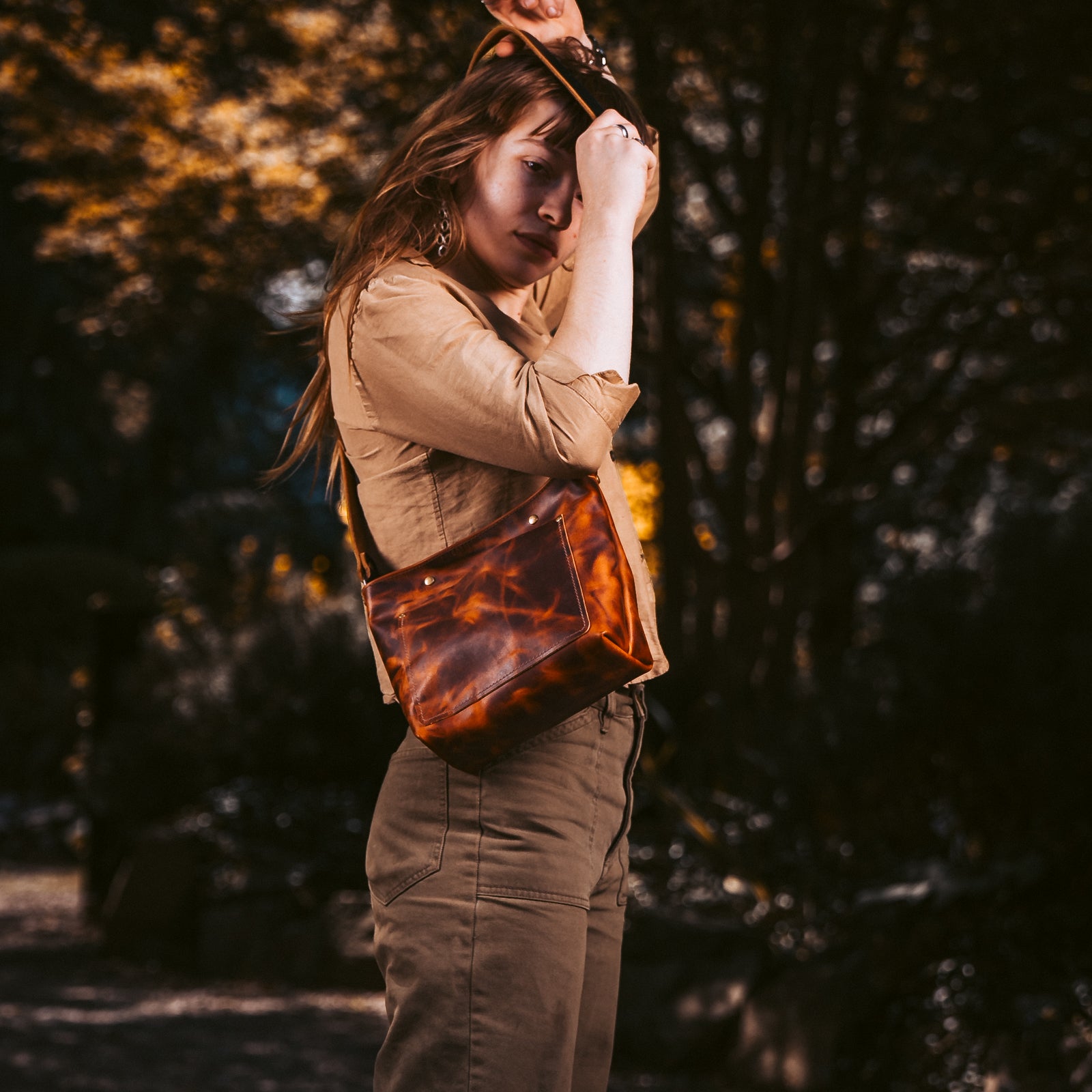 A woman wearing a tan shirt and brown cargo pants carries a brown tortoiseshell leather shoulder bag in an outdoor forest setting.