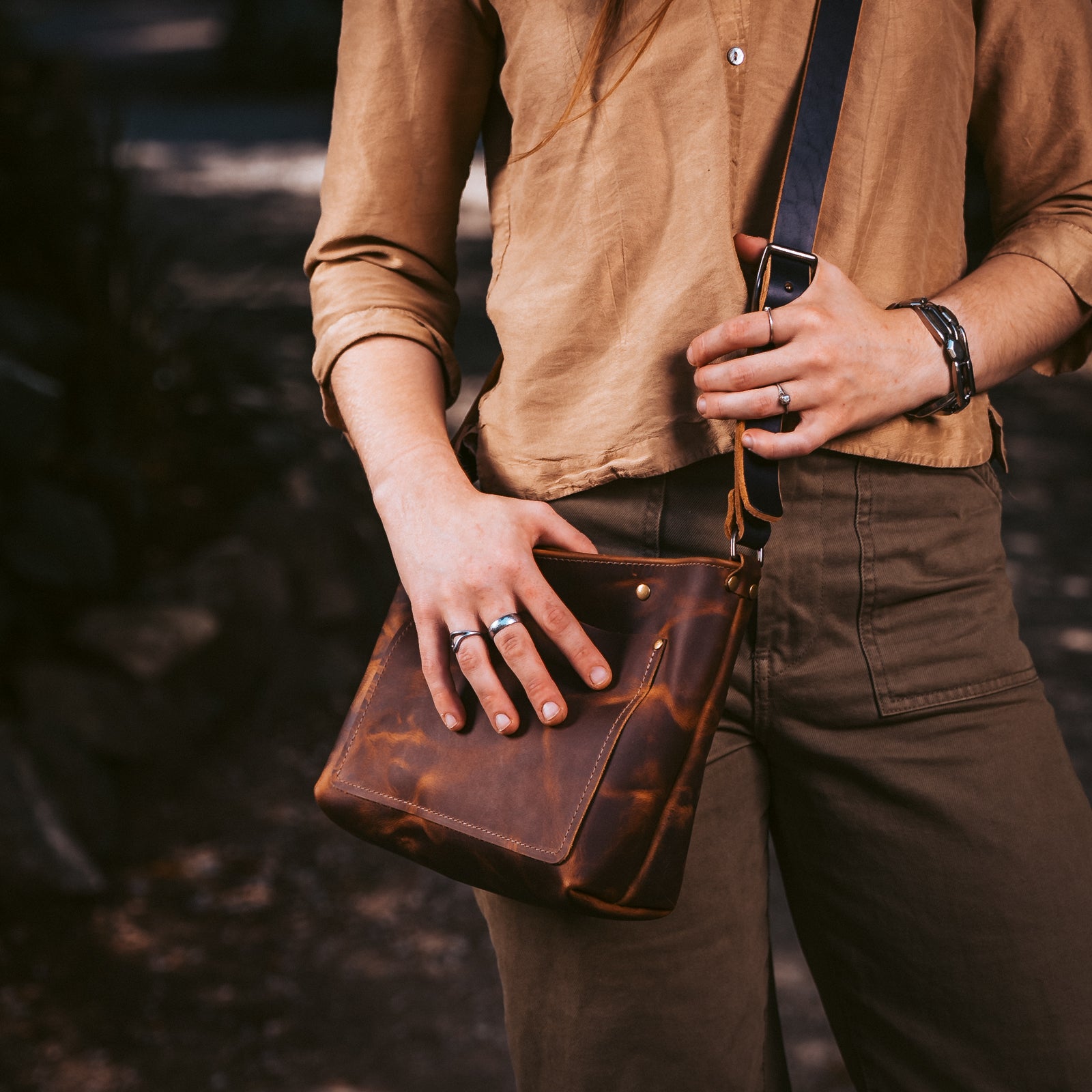 A person wearing a tan shirt and dark pants holds a brown leather crossbody bag with a dark strap in an outdoor setting.